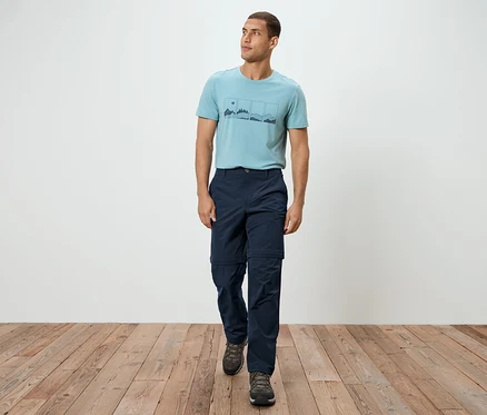 Un homme marche sur un plancher en bois, il porte un t-shirt bleu, un pantalon bleu marine et des baskets grises.