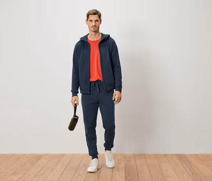 Un homme debout en survêtement bleu marine et t-shirt rouge tenant une bouteille d'eau.