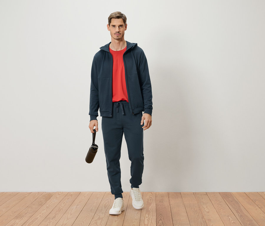 Un homme debout en survêtement bleu marine et t-shirt rouge tenant une bouteille d'eau.