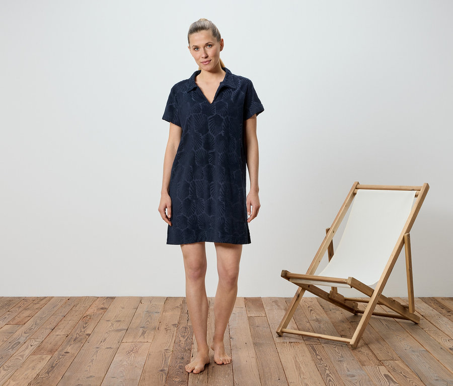 Une femme se tient pieds nus sur un plancher en bois devant un fond blanc. Elle porte une robe bleu marine à manches courtes. Sur sa droite se trouve une chaise longue en bois avec un tissu blanc.