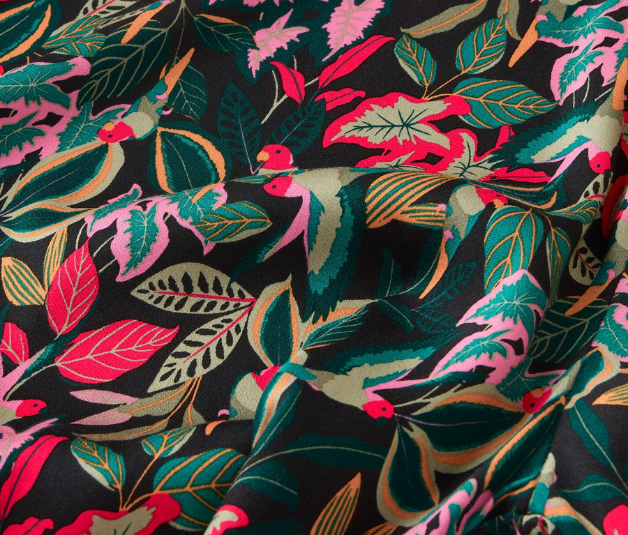 Gros plan d'un tissu avec un motif de feuilles tropicales et d'oiseaux dans des tons de vert, rose et rouge sur un fond noir.