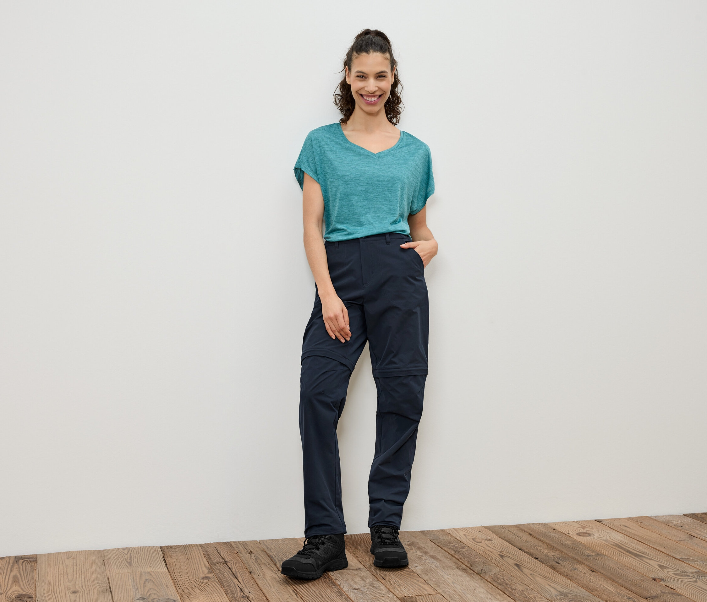 Une femme se tient devant un mur blanc et porte un t-shirt bleu-vert, un pantalon zippé bleu marine et des chaussures noires.