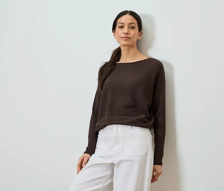 Femme appuyée contre un mur portant un pull marron et un pantalon blanc.