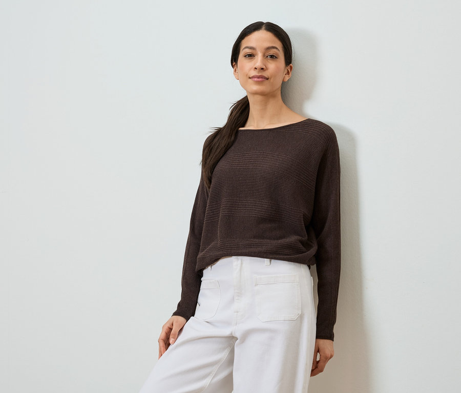 Femme appuyée contre un mur portant un pull marron et un pantalon blanc.