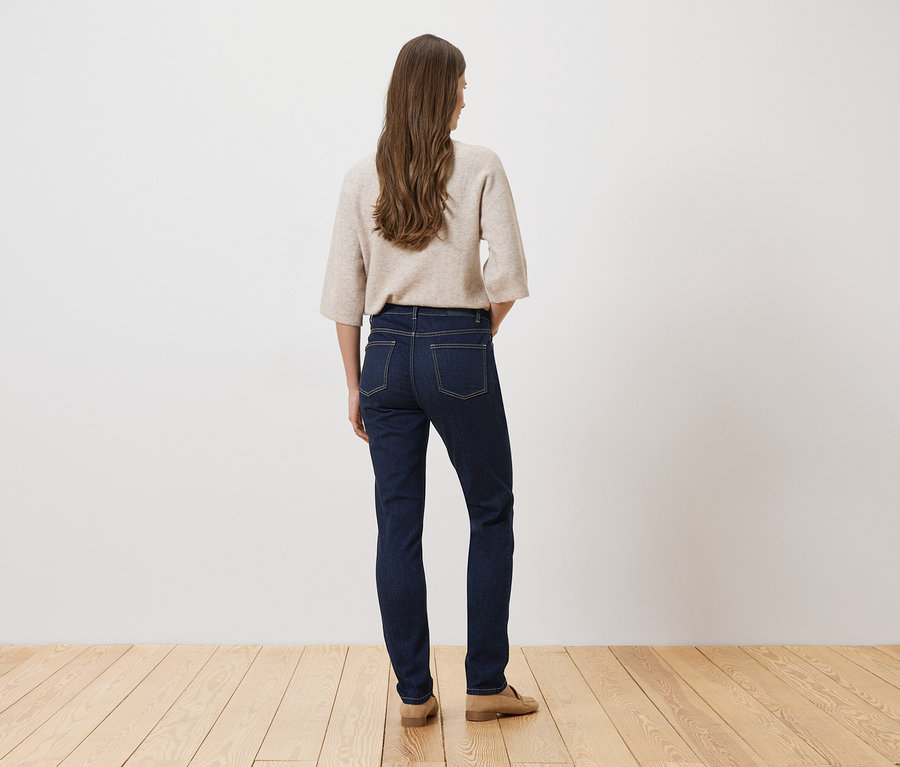 Une femme aux longs cheveux bruns se tient dos à la caméra, portant un jean foncé, un pull beige et des chaussures beiges.