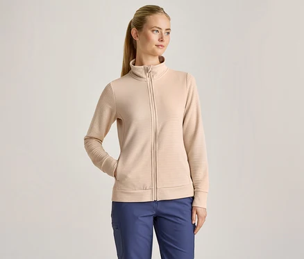 Une femme aux cheveux blonds attachés en queue de cheval porte une veste zippée côtelée beige et un pantalon bleu, une main dans la poche de la veste.