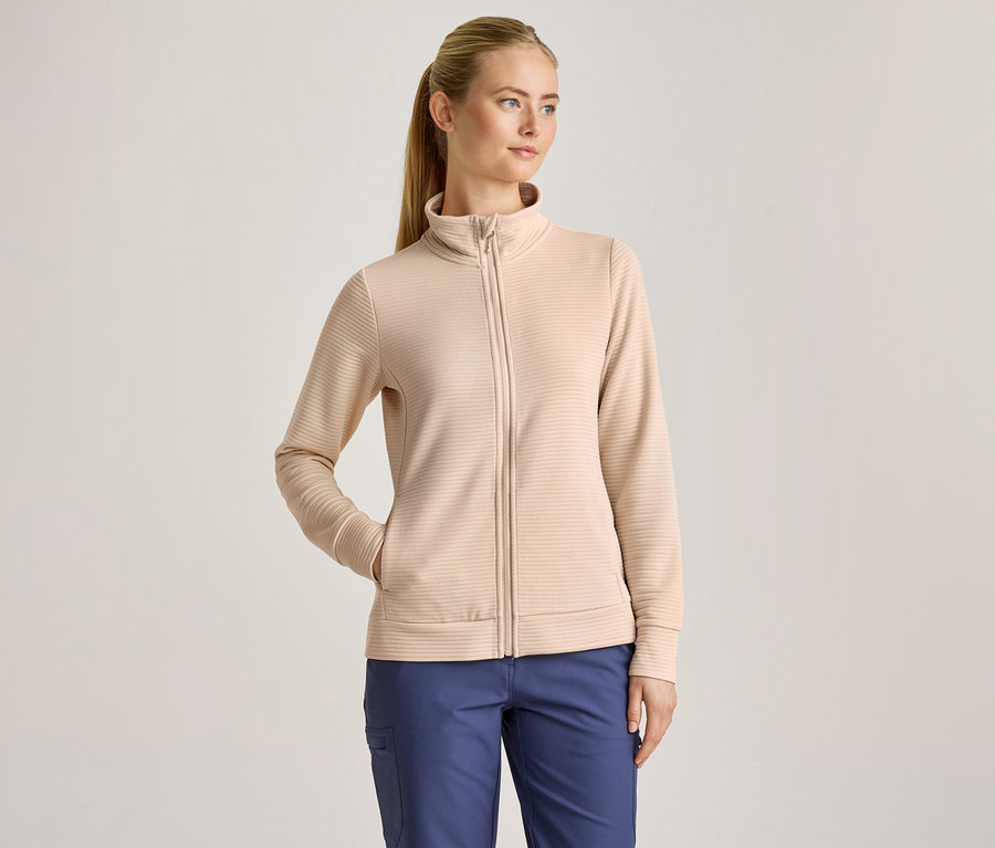 Une femme aux cheveux blonds attachés en queue de cheval porte une veste zippée côtelée beige et un pantalon bleu, une main dans la poche de la veste.
