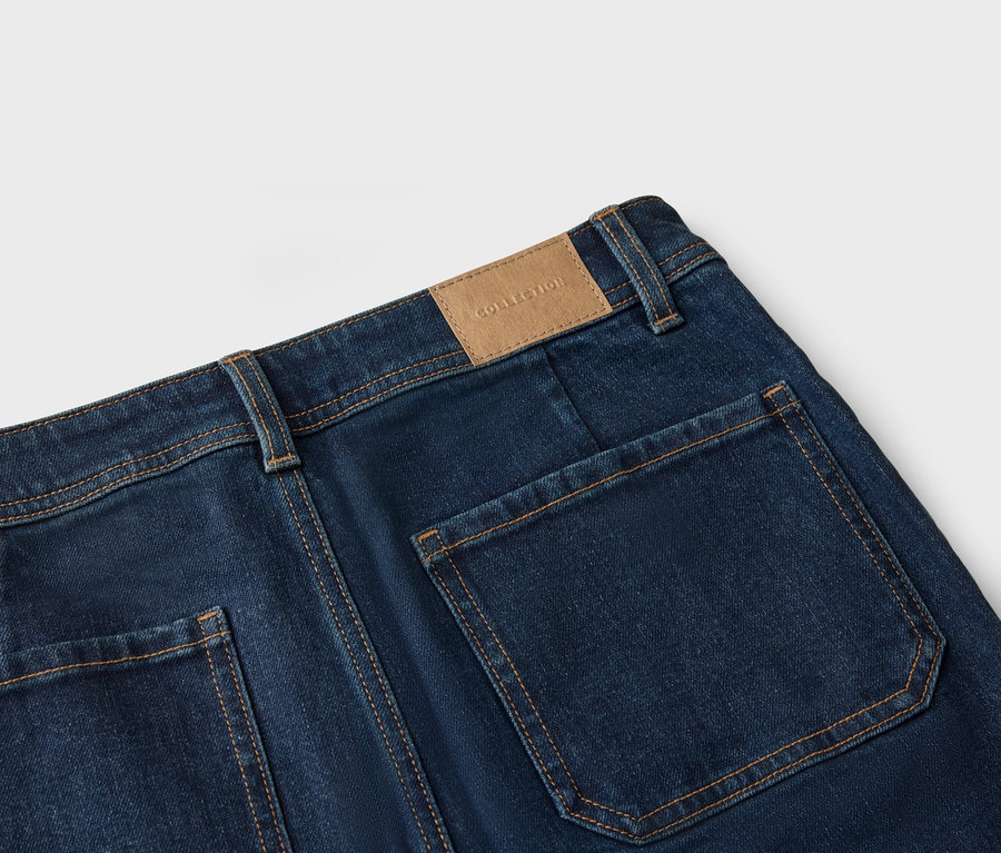 Gros plan sur l'arrière d'un jean bleu foncé avec des poches plaquées et un patch en cuir marron avec l'inscription « Collection ».