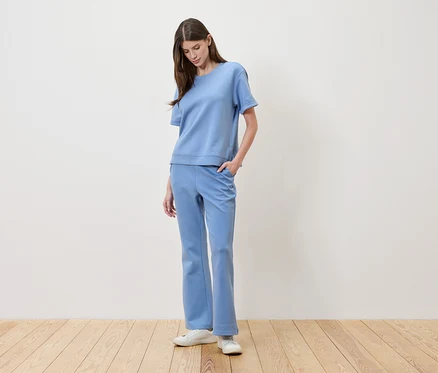 Une femme aux cheveux bruns porte un t-shirt bleu clair et un pantalon assorti, avec une main dans la poche.