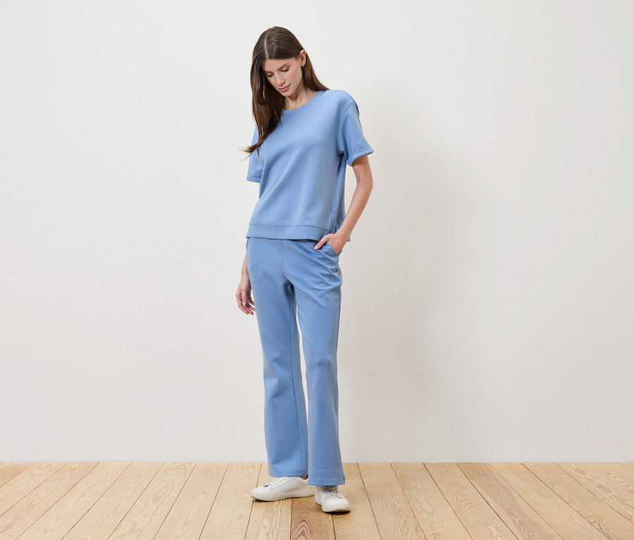 Une femme aux cheveux bruns porte un t-shirt bleu clair et un pantalon assorti, avec une main dans la poche.