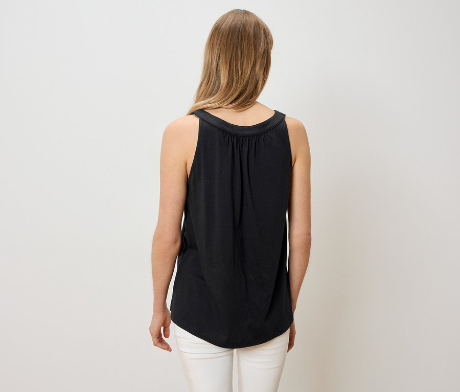 Vue de dos d'une femme aux longs cheveux blonds, portant un haut noir sans manches et un pantalon blanc.
