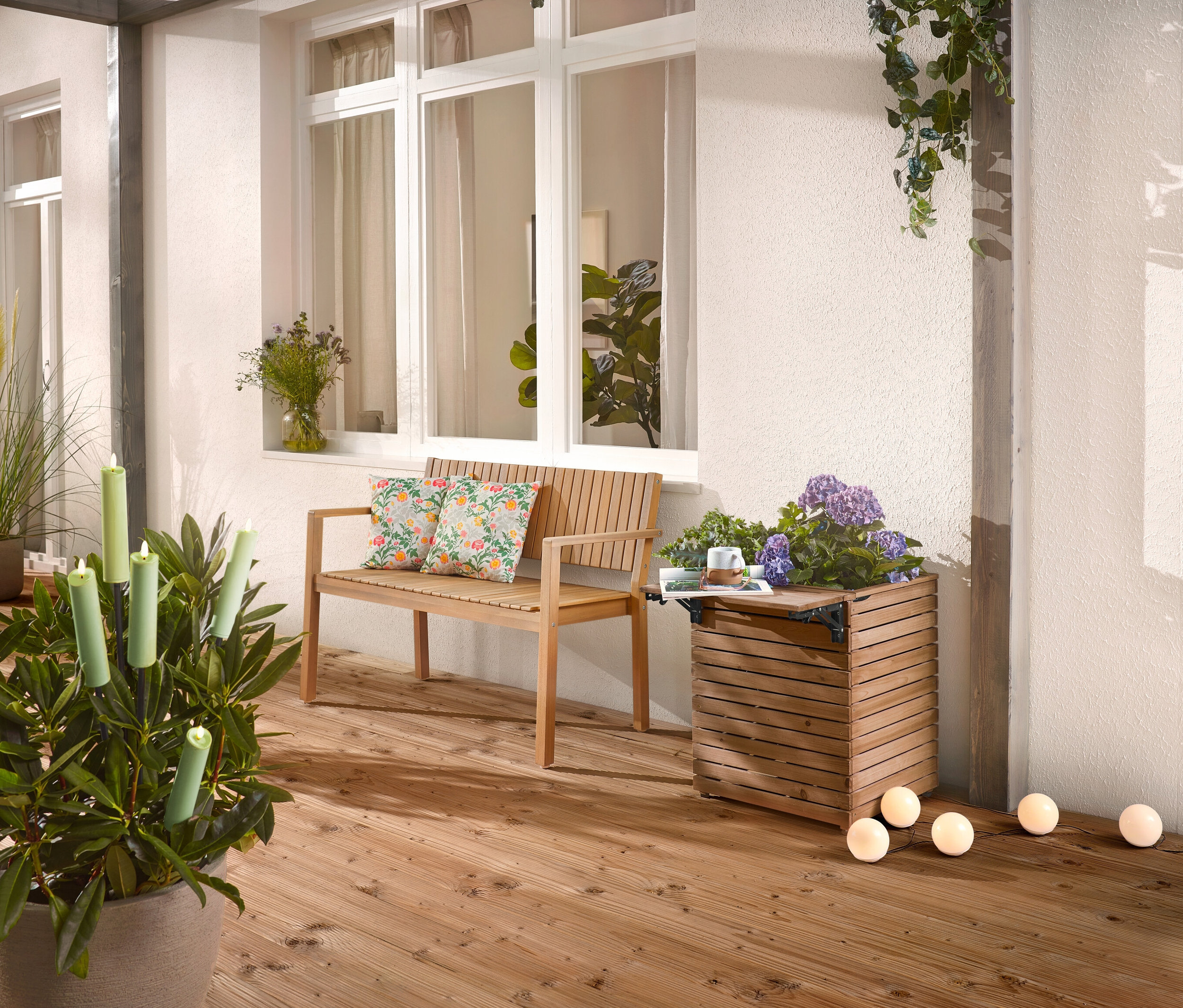 Sur une terrasse en bois se trouvent un banc en bois avec des coussins, un bac à plantes avec des fleurs et une plante en pot avec des bougies.