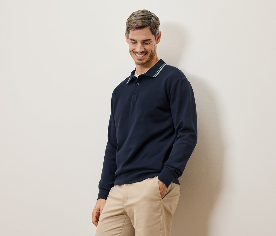 Un homme souriant portant un polo à manches longues bleu marine et un chino beige se tient devant un mur couleur crème.
