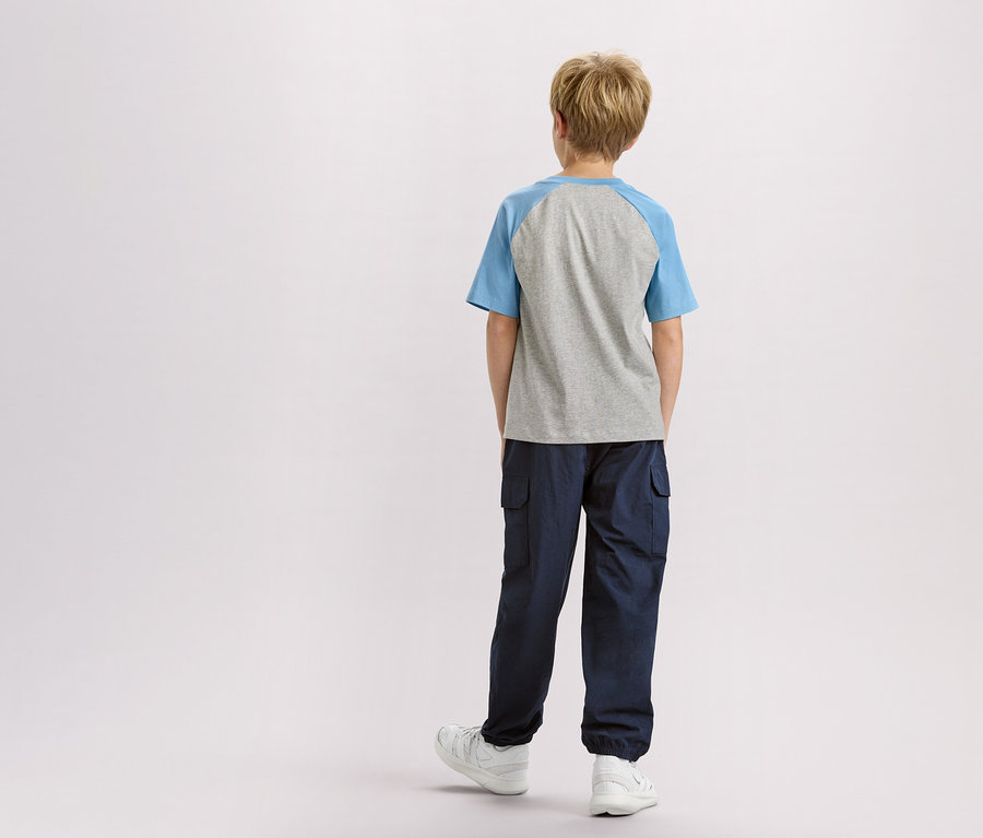 Vue de dos d'un enfant blond portant un t-shirt gris à manches bleues, un pantalon bleu marine et des baskets blanches.