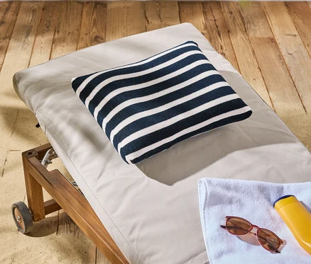 Un coussin rayé repose sur une chaise longue à roulettes à côté d'une crème solaire, de lunettes de soleil et d'une serviette.