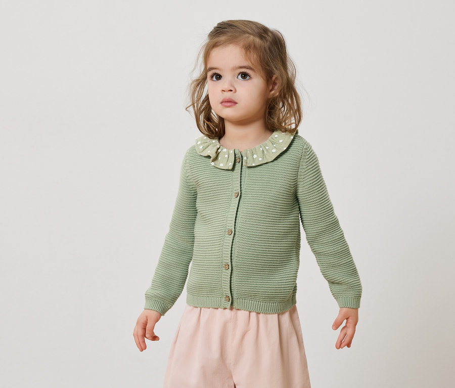 Une petite fille porte un cardigan en tricot vert et une jupe rose.