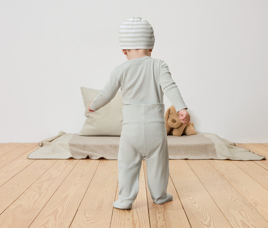 Un bébé en pyjama vert clair et bonnet rayé se tient dos à la caméra sur un plancher en bois.