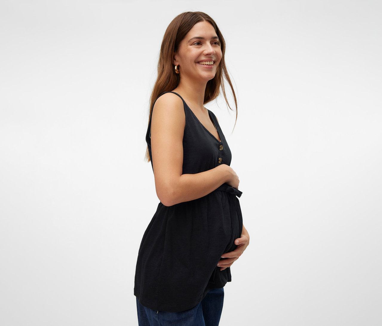 Une femme souriante tient son ventre, portant un top d’allaitement noir MAMALICIOUS « MLEVI LIA » et un jean.