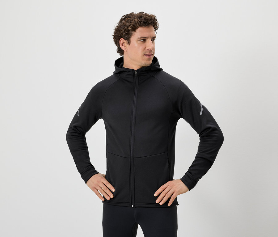 Homme aux cheveux bouclés portant un sweat à capuche noir zippé et un legging noir, les mains sur les hanches.