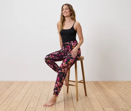 Une femme est assise pieds nus sur un tabouret, portant un débardeur noir et un pantalon à fleurs.