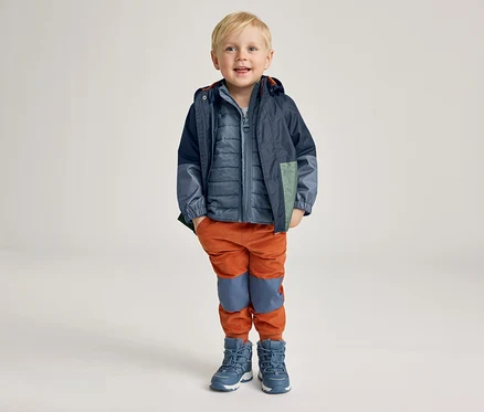 Un garçon souriant aux cheveux blonds courts porte une veste bleue, un gilet bleu matelassé, un pantalon orange et des bottes bleues.