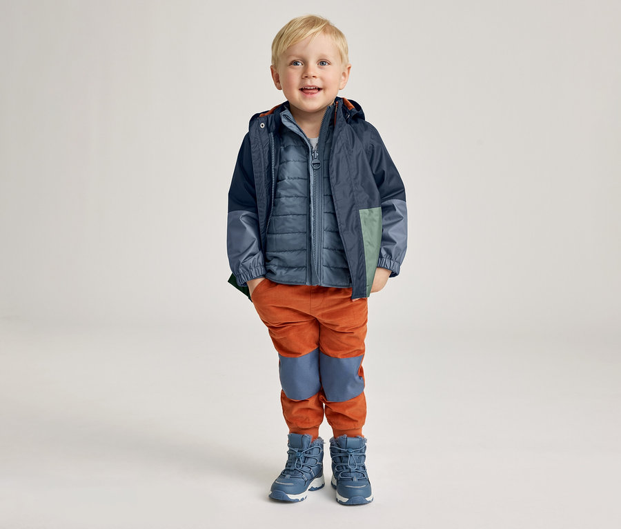 Un garçon souriant aux cheveux blonds courts porte une veste bleue, un gilet bleu matelassé, un pantalon orange et des bottes bleues.