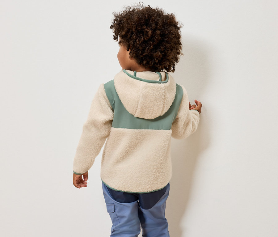 Vue arrière d'un enfant aux cheveux bouclés portant une veste polaire crème à capuche avec des accents verts et un pantalon bleu.
