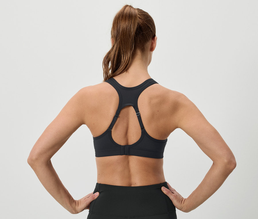 Vue de dos d'une femme aux cheveux bruns relevés en queue de cheval, portant un soutien-gorge de sport noir et un legging, les mains sur les hanches.