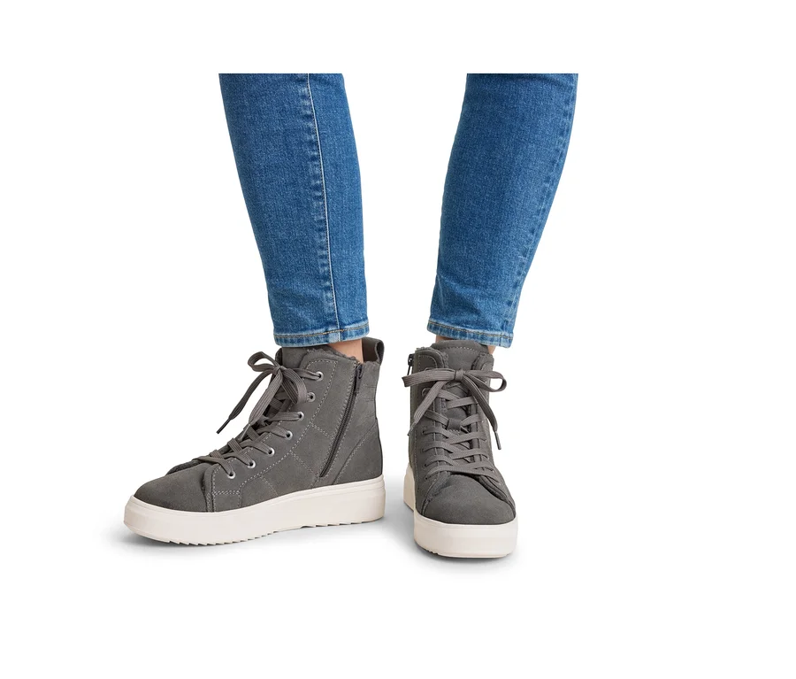 Jambes d'une personne portant un jean bleu et des baskets montantes grises avec des semelles blanches, des lacets et des fermetures éclair latérales.