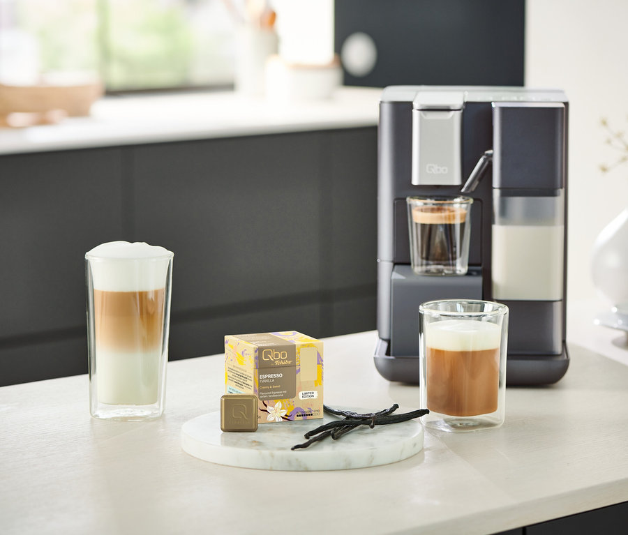 Un Qbo MILK MASTER Gentle Grey est placé à côté de deux verres de café. Un Qbo MILK MASTER rouge est visible en arrière-plan.