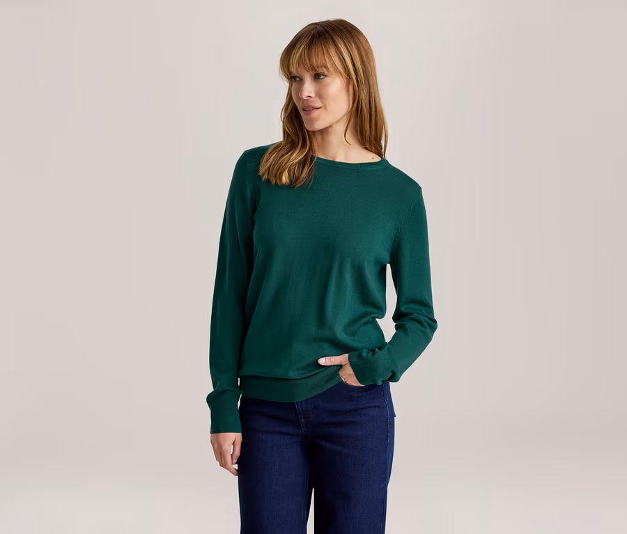Femme portant un haut vert à manches longues et un jean bleu foncé, la main droite dans la poche, regardant vers la droite.