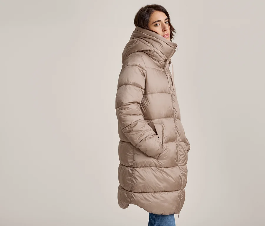 Femme portant un long manteau matelassé beige avec un col montant et une capuche, les mains dans les poches, regardant la caméra.