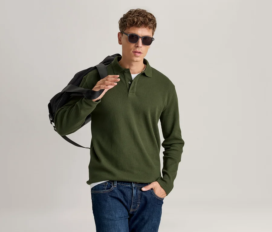 Homme portant un polo vert à manches longues, un jean bleu et des lunettes de soleil, avec un sac noir sur l'épaule.