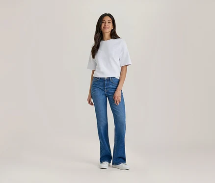 Une femme aux longs cheveux foncés, vêtue d'un haut blanc à manches courtes et d'un jean bleu évasé, se tient de face et sourit. Elle porte également des baskets blanches.