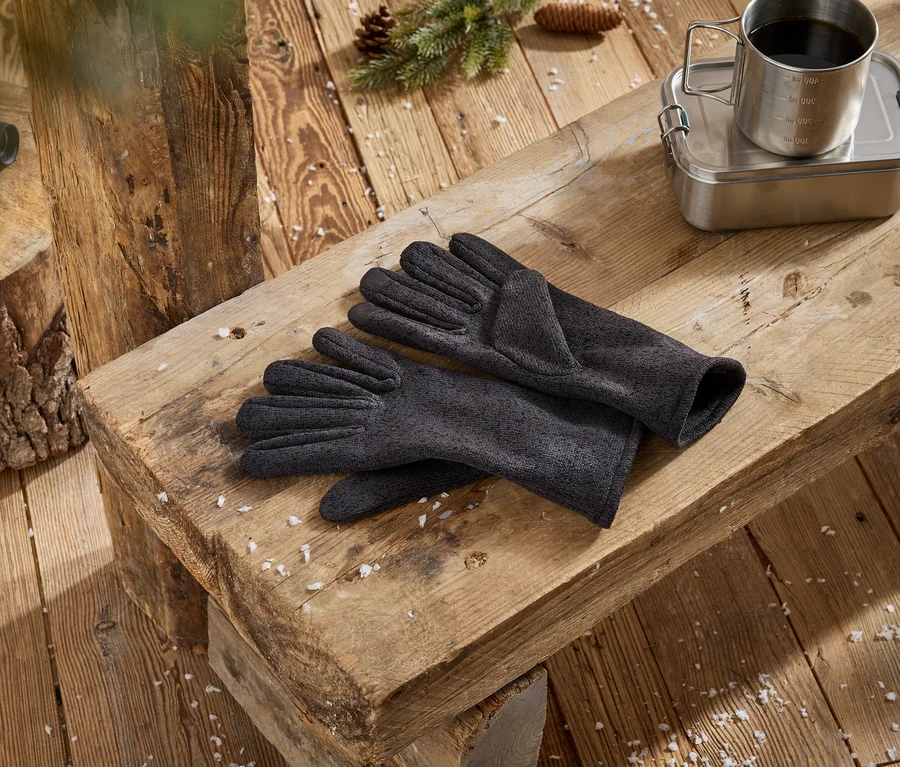 Une paire de gants gris foncé repose sur un banc en bois rustique, à côté d'une boîte à lunch en métal avec une tasse de café en métal dessus. Des jumelles noires sont posées sur une souche d'arbre en bois à gauche, avec des pommes de pin et de la neige éparse sur la surface en bois.
