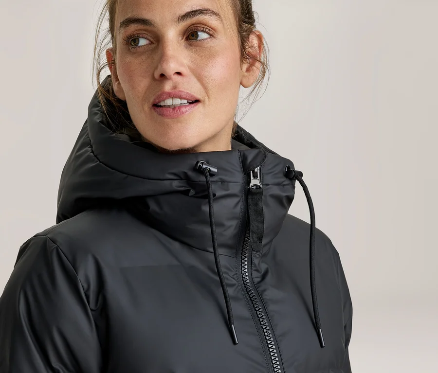 Femme portant une veste noire à capuche avec fermeture éclair et cordons de serrage, regardant vers la droite.