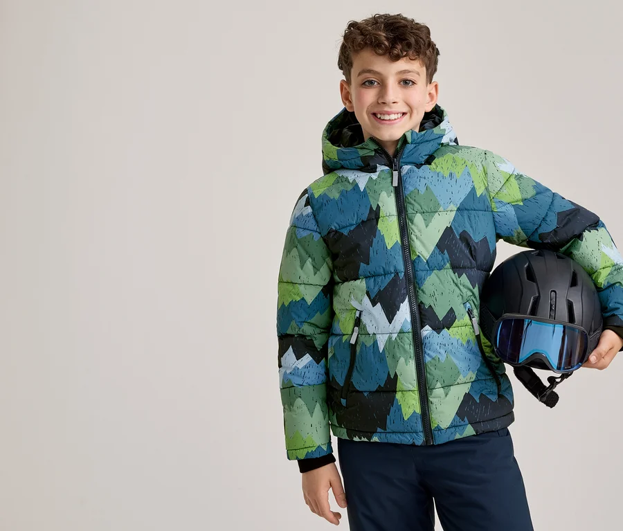 Un jeune garçon souriant vêtu d'une veste d'hiver à motifs verts, bleus et noirs tient un casque de ski noir avec des lunettes bleues.