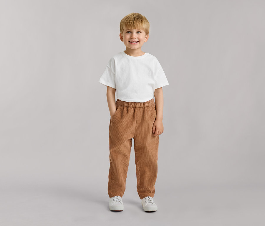 Jeune garçon blond souriant, portant un t-shirt blanc, un pantalon en velours côtelé marron et des baskets blanches, les mains dans les poches.