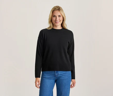 Femme blonde souriante portant un pull noir à manches longues et un jean bleu, debout devant un fond clair.