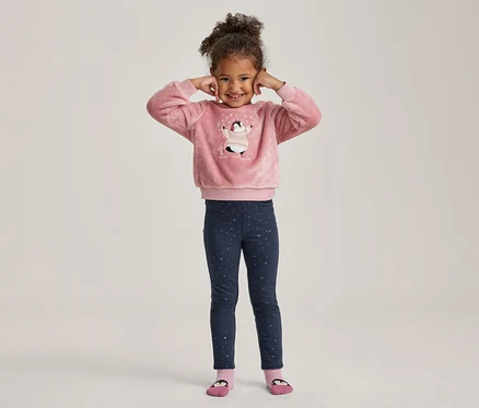 Une petite fille souriante aux cheveux bouclés, les mains aux oreilles, porte un haut rose duveteux avec un motif de pingouin et un legging bleu foncé à étoiles.