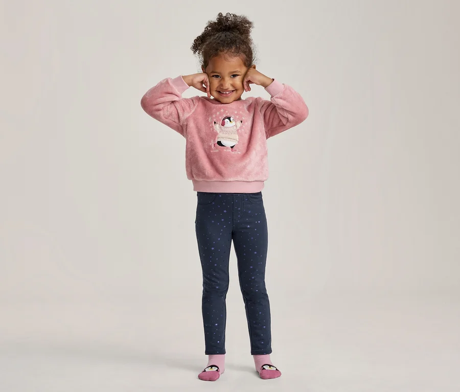 Une petite fille souriante aux cheveux bouclés, les mains aux oreilles, porte un haut rose duveteux avec un motif de pingouin et un legging bleu foncé à étoiles.