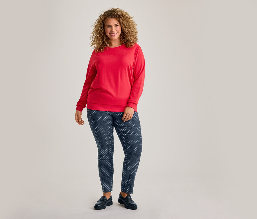 Femme souriante aux cheveux bouclés, portant un haut rouge à manches longues, un pantalon bleu marine à pois blancs et des chaussures noires.