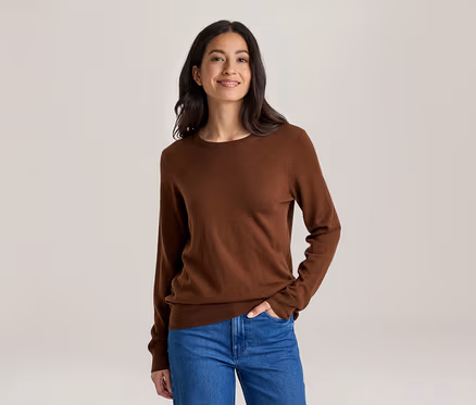 Une femme souriante aux cheveux foncés porte un haut marron à manches longues et un jean bleu, une main dans la poche, sur un fond clair.