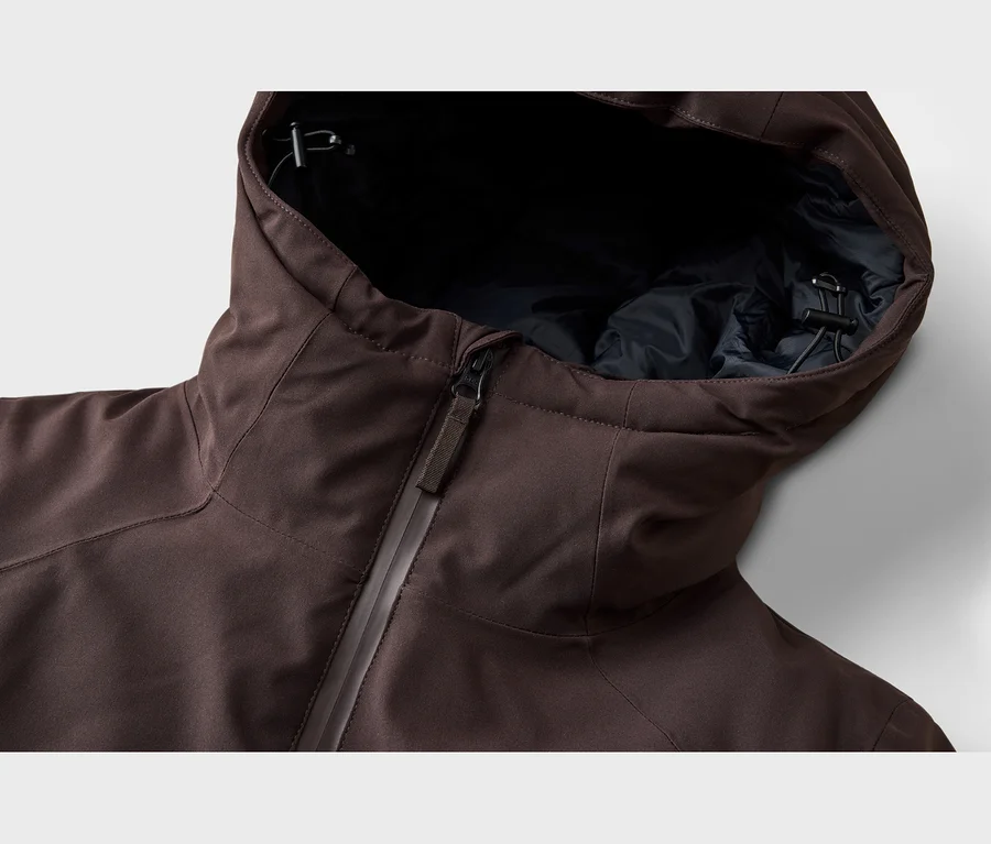 Gros plan d'une veste marron avec une fermeture éclair partiellement ouverte, révélant une doublure intérieure foncée. La capuche est relevée, montrant un cordon de serrage noir avec un stoppeur.