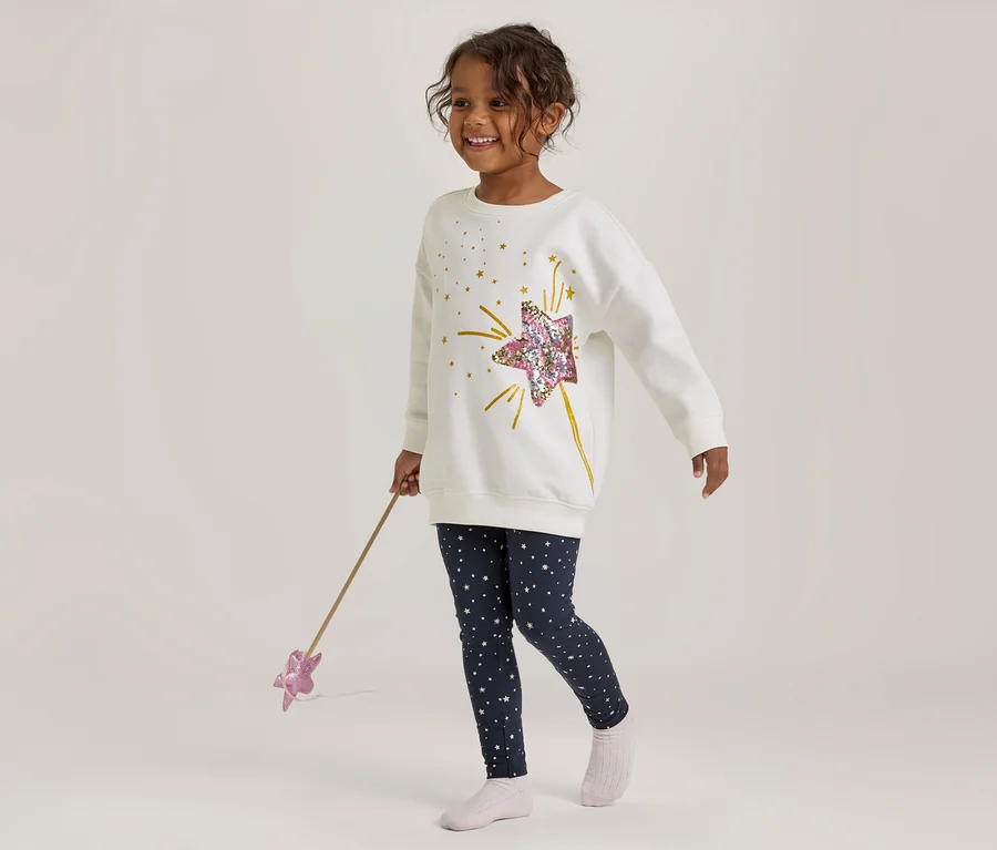 Fille souriante portant un haut blanc à manches longues avec une étoile à sequins et un legging bleu marine à étoiles, tenant une baguette rose.