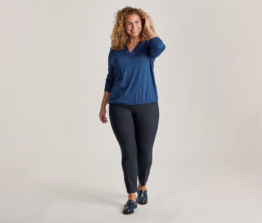 Une femme souriante aux cheveux bouclés avance, vêtue d'un haut bleu à motifs avec col en V, d'un pantalon slim foncé et de mocassins foncés, une main dans ses cheveux.