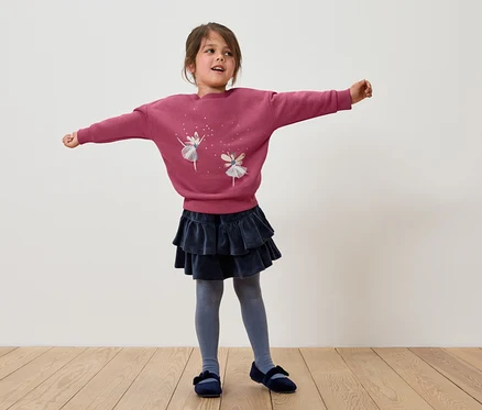 Une fillette se tient debout, les bras écartés, portant un sweat-shirt rose avec des ballerines, une jupe bleu foncé à volants, des collants gris et des chaussures bleues.