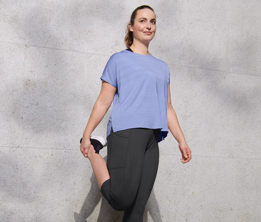 Femme étire sa jambe en portant un corsaire de sport 3/4 et un t-shirt de sport violet chiné.