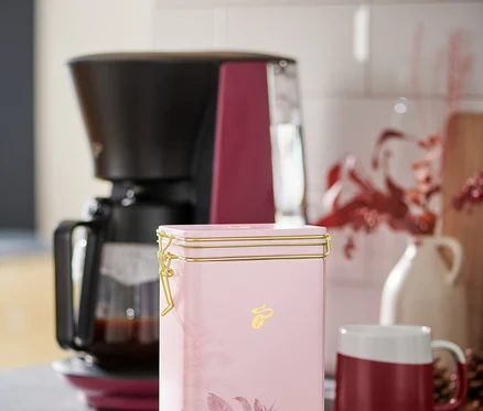 Une boîte à café rose est posée sur un comptoir de cuisine à côté d'une cafetière et d'une tasse rouge et blanche.