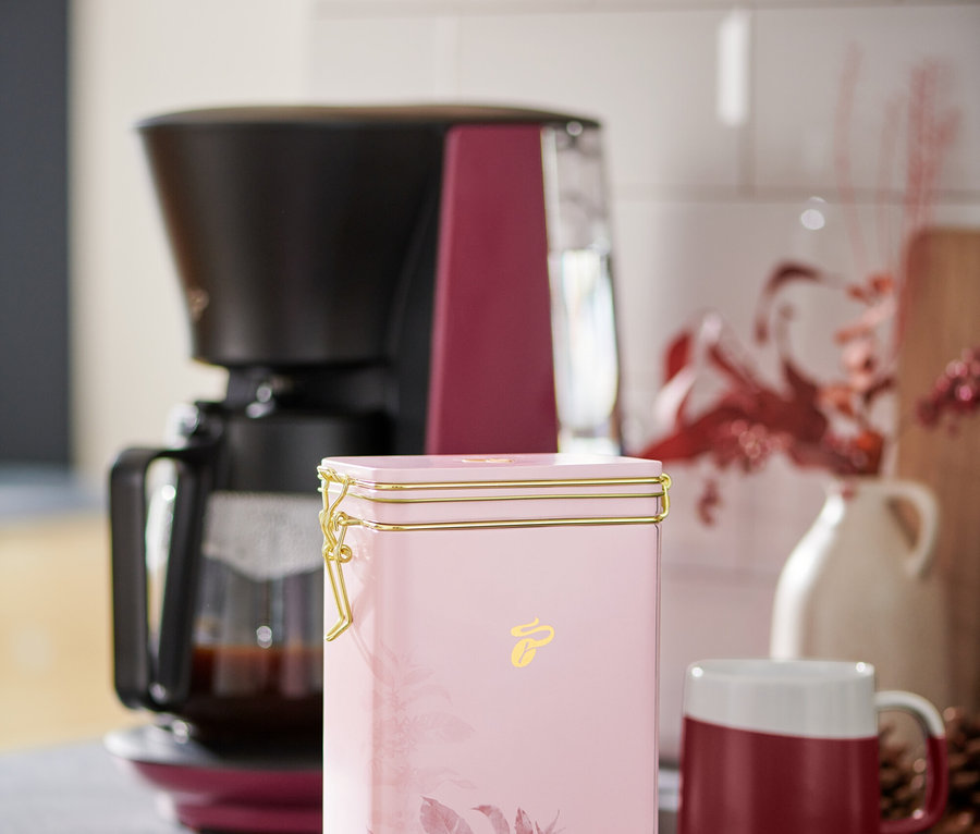 Une boîte à café rose est posée sur un comptoir de cuisine à côté d'une cafetière et d'une tasse rouge et blanche.